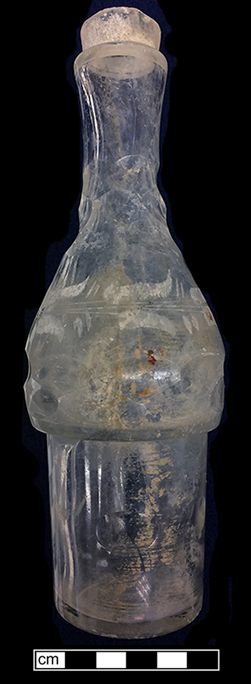 Colorless glass caster. Vessel has cut ovals along the base, body and neck, as well as engraved .  Floral and leaf decoration on the body.  The finish is ground on the exterior of the lip, suggesting that it once had a perforated metal top and was used for sugar or pepper. The narrow lower body of this vessel indicates that it was part of a set that was stored in a metal stand. 51NW262.
