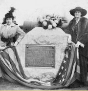 Figure 3 Black and white historical photograph showing two women in early 20th century dress and wide-brimmed hats standing beside a memorial topped with white flowers and draped with American flags, with a commemorative plaque mounted on the stone.
