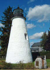 Figure 4 White-painted conical lighthouse with black lantern room and railing at top, commemorative monument in foreground.