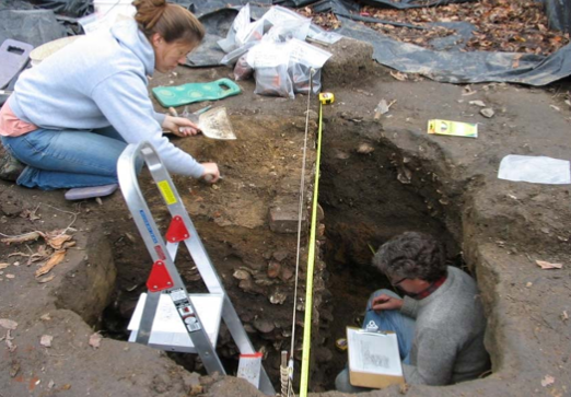A man crouched in a deep hole with a clipboard and a woman kneeling near the hole with a trowel in her hand.