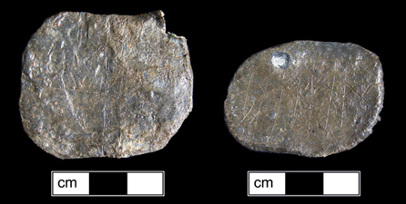Two side-by-side flat oval objects of lead with grids and x-s scratched on them.