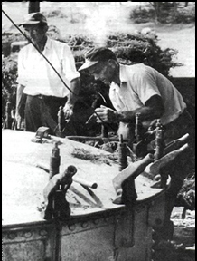 Figure 2 Black and white historical photograph showing two men in work clothes stand beside industrial equipment, one closely inspecting or working on the machinery outdoors.