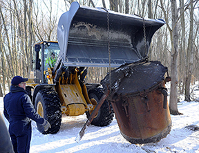Figure 4 A yellow excavator in a snowy winter forest, with chains securing a heavily corroded cylindrical artifact as museum staff in winter clothing supervise the recovery operation.