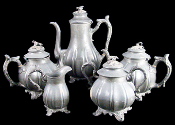 Figure 1 Five-piece Britannia metal tea and coffee service set displayed against black background, featuring ornate fluted bodies with decorative handles, spouts, and animal finials on lids, 
                    showing varying heights from creamer to tall coffeepot.