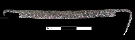 Figure 1 Archaeologically recovered drawknife against black background with 5 cm scale bar, showing the curved metal blade structure and tangs for handles.
