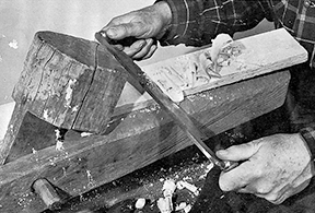 Figure 3 Black and white photograph showing hands using a drawknife on a shaving horse, with wood shavings accumulating as the blade removes material from a wooden piece secured in a traditional woodworking jig.