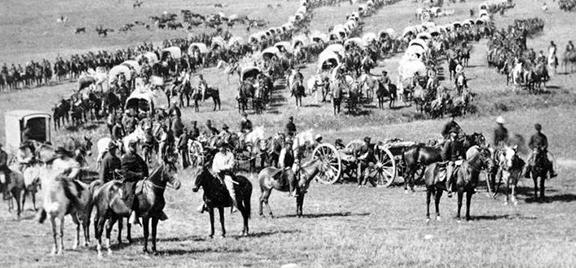 Figure 3 Historical black and white photograph showing a long wagon train stretching across an open plain, with numerous covered wagons, horses, riders, and people forming a caravan 
                    migration scene typical of westward expansion.