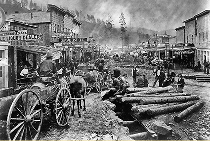 Figure 5 Historical black and white photograph of a frontier town main street with wooden storefronts including a liquor dealer and bank, showing horse-drawn wagons, groups of people, 
                    and large logs laid across the muddy street, with forested hillside and smoke in the background.