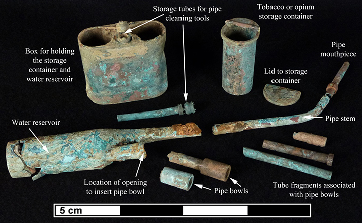 Figure 1 Archaeological assemblage of corroded Chinese opium smoking paraphernalia with labeled components against black background, including rectangular storage box with turquoise patina, 
                    cylindrical tobacco container, pipe stems and bowls, water reservoir, cleaning tools, and various tube fragments, with 5 cm scale bar.
