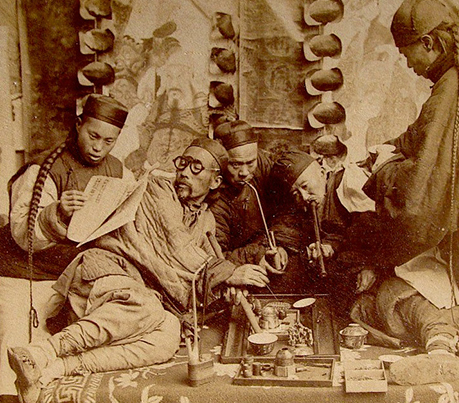 Figure 4 Sepia-toned historical photograph showing a group of Chinese men in traditional dress seated around a low table with opium smoking equipment, including long pipes, with decorative 
                    hanging in the background.