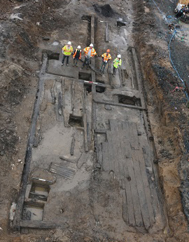 Figure 1 Aerial view of fully exposed archaeological foundations of a building showing preserved wooden floor joists with interior floor planking visible in waterlogged sediment, with team of archaeologists in safety gear standing along edge for scale.