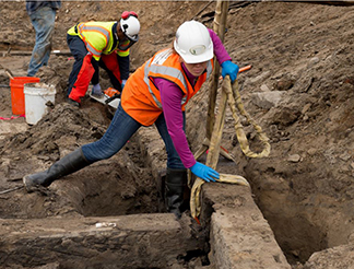 Figure 2 Conservator in orange safety vest, white hard hat and blue gloves kneeling in excavation trench while attaching lifting straps to deteriorated wooden foundation timbers, with another worker visible in background.