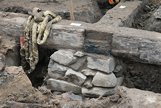 Figure 3 Close-up view of partially excavated archaeological wooden foundation structure showing weathered timber beams joined together with stone pier visible beneath.