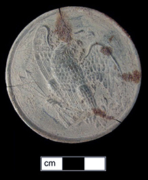 Archaeological Civil War era round scabbard belt plate with heavily corroded green-gray patina against black background with centimeter scale bar, showing faint remnants of embossed eagle motif.