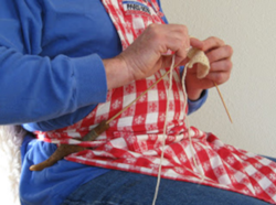Close-up photograph of hands working on knitting project with wooden knitting needle and sheath tucked into a red and white gingham checkered apron.