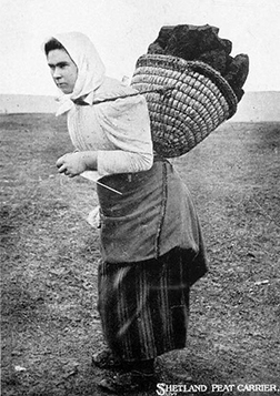 Historical black and white photograph captioned showing woman in traditional dress with headscarf and long skirts knitting using a needle sheath while carrying large woven basket filled with dark peat blocks on her back across barren landscape.