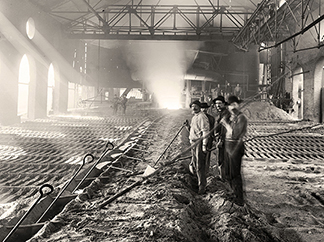 Historical black and white photograph of industrial foundry interior showing workers standing among rows of pig iron on floor in smoky atmospheric conditions.