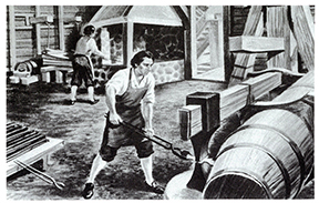 Historical engraving showing industrial worker in apron using long-handled tool to tend large cylindrical furnace flames, with other workers visible in background of foundry setting.