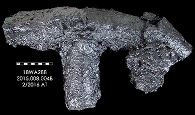 Photograph of corroded T-shaped pig iron fragment against black background, with identification label 