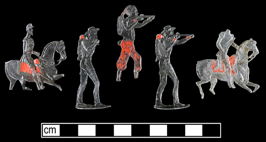 Five lead toy soldiers with traces of red paint on a black background with a scale bar. 