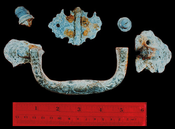 Corroded metal coffin furniture handle in five fragments on a black background with a scale.