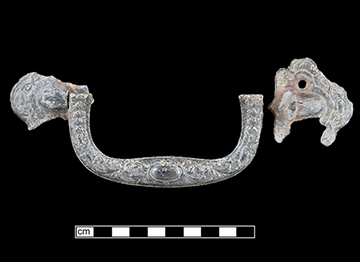 Metal coffin furniture handle in two fragments on a black background with a scale.