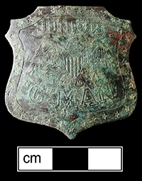 Corroded shield-shaped metal badge photographed against a black background with a centimeter scale bar. The surface shows corrosion products obscuring most details, with barely visible embossed text and the eagle design showing through the corrosion layers