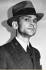 Black and white portrait photograph of a young man wearing a dark suit and fedora hat, looking directly at the camera with a serious expression.