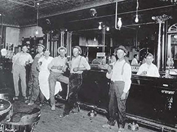 Seven men standing at a bar in an early 20th-century saloon.