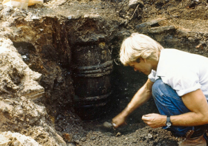 Color photograph of an archaeologist in a white shirt and jeans kneeling at the edge of an excavation unit, carefully exposing a wooden barrel in situ. The barrel remains partially buried in dark soil.