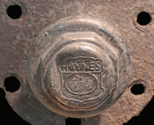 Close-up photograph of a corroded hub cap with mounting holes. The surface shows a circular embossed design in the center with text reading 'HAYNES'