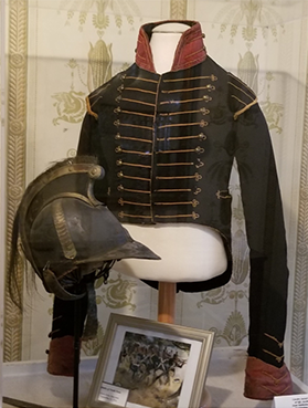 A museum display of the helmet and related items including a coatee (looks like a coat)
mounted on a mannequin. The coatee is black or navy with gold-colored shoulder
details and 26 gold-colored buttons with prominent gold-colored stitching. A framed
photo sits on the display shelf below the coatee.