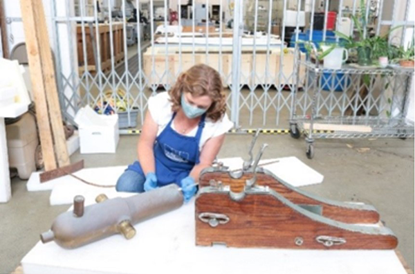 Conservator working on a Lyle gun and its wood and metal stand in the
conservation lab. The gun is about two feet long and made of copper alloy.