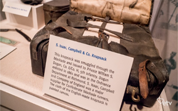 Museum display of a knapsack including a museum label that reads “5. Isaac,
Campbell & Co. Knapsack. This knapsack was smuggled through the blockade and
issued to Private William S. Dogan, Co E/Co. H 5 th infantry. Dogan enlisted in 1861 and
was in the Ordnance Department at Richmond. S. Isaac Campbell and Company of
England was a major supplier for Confederate blockade runners. The cloth of the
English-made knapsack is painted canvas.”