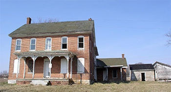 Photo of a two-story farmhouse with large porch with extension off the back of the house. Two whitewashed wood outbuildings in the background.
