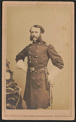Historic sepia-tone photo of a Caucasian man in military uniform with large white gloves. The man has short curly hair, prominent sideburns, and a mustache.