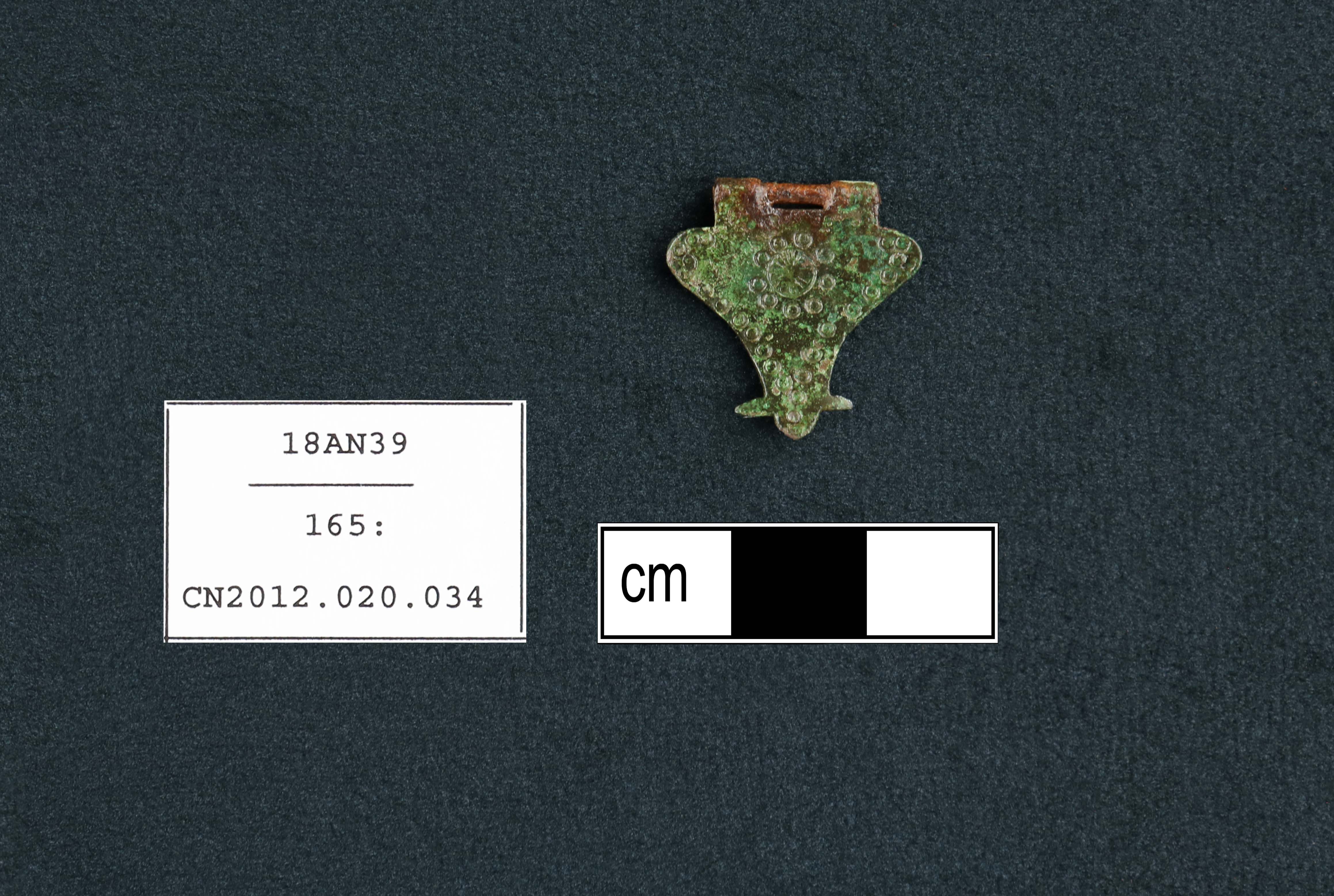 Decorative triangular-shaped copper alloy artifact with corroded surface. The photo scale shows the artifact is less than 3 cm wide.