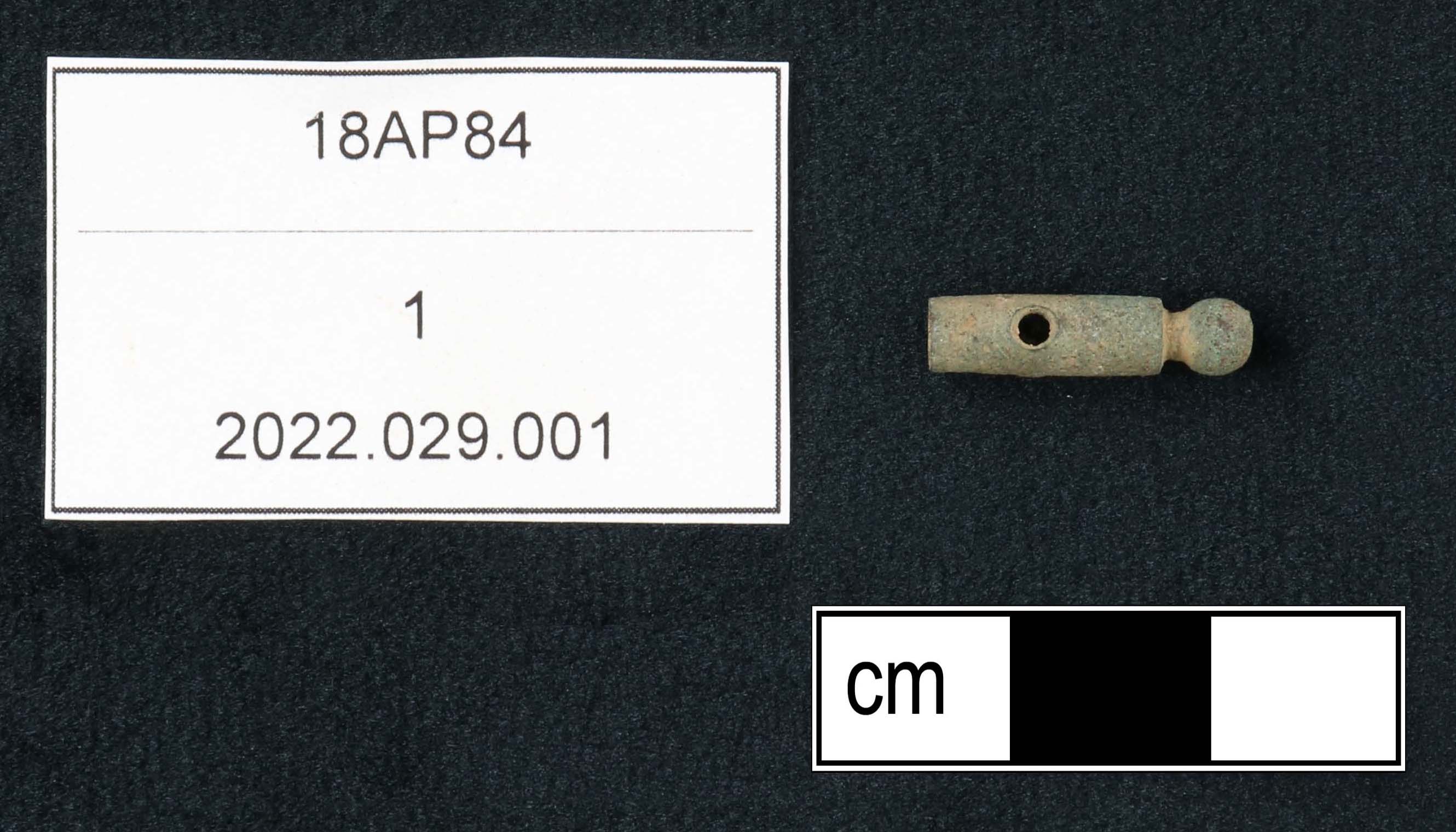 Very small copper alloy artifact that is no longer than 1.5 centimeters. A rounded ball-tip end attaches to a narrow cylinder two times the length of the ball-tip end. A drilled hole is bored through the side of the cylinder.