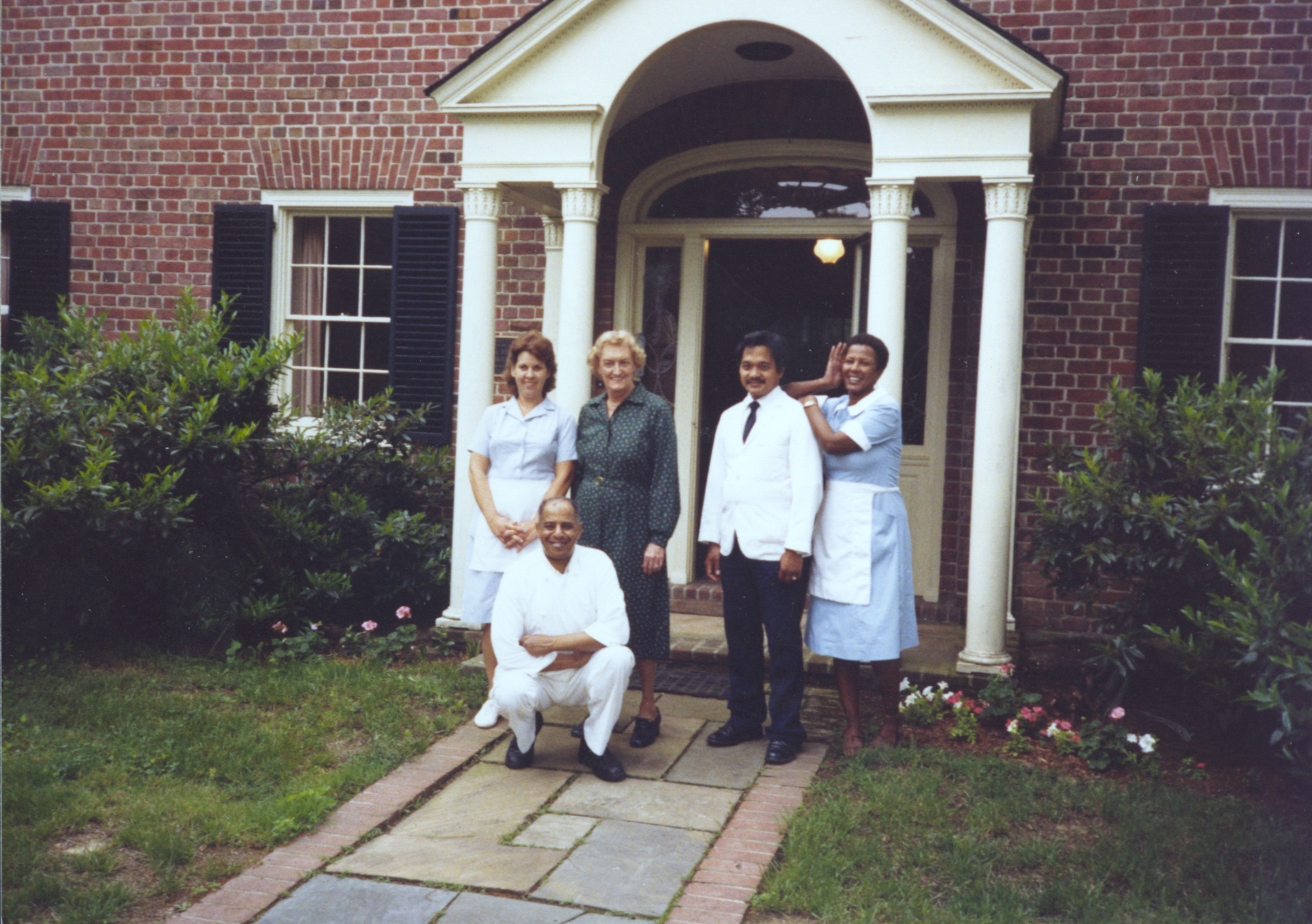 Photograph of group of individuals in front of front door of brick manor house. Person to the far right is wearing a maid's uniform.