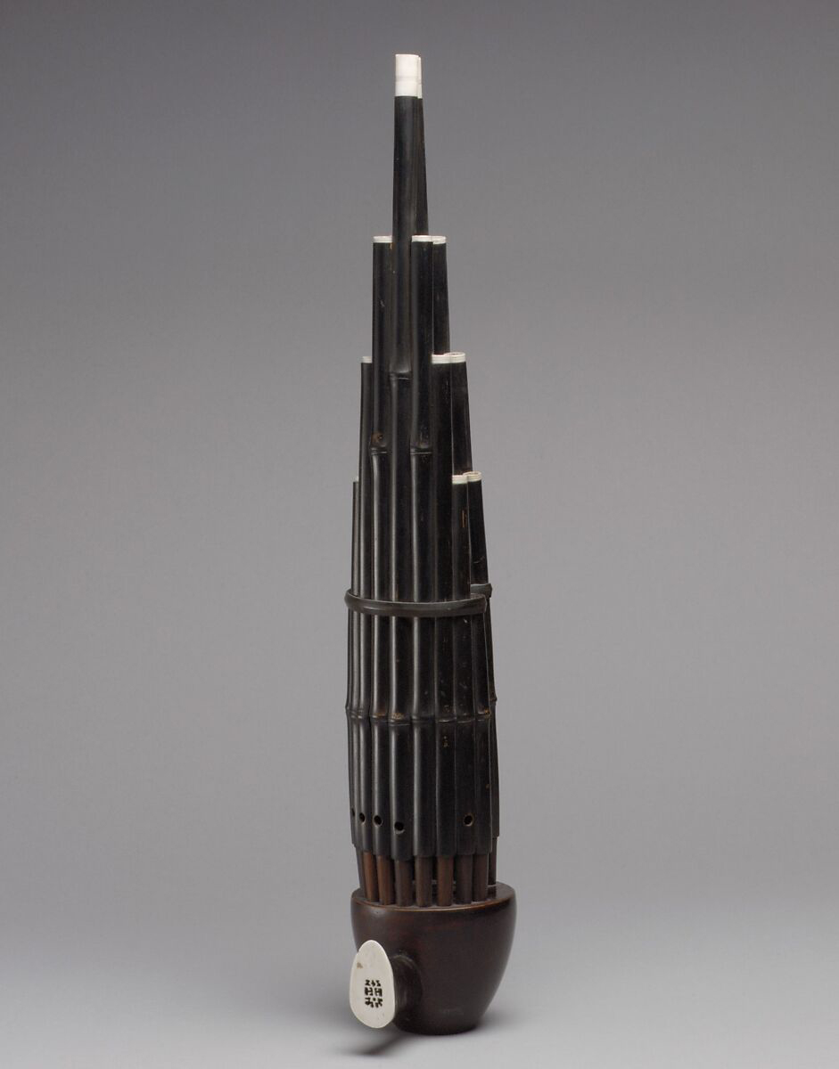 An instrument shaped like a bundle of bamboo cut to different lengths. One mouthpiece at the base opens to a small cup-shaped wind chamber. Holes in the sides of the bamboo-shaped pieces indicate a noisemaker.
