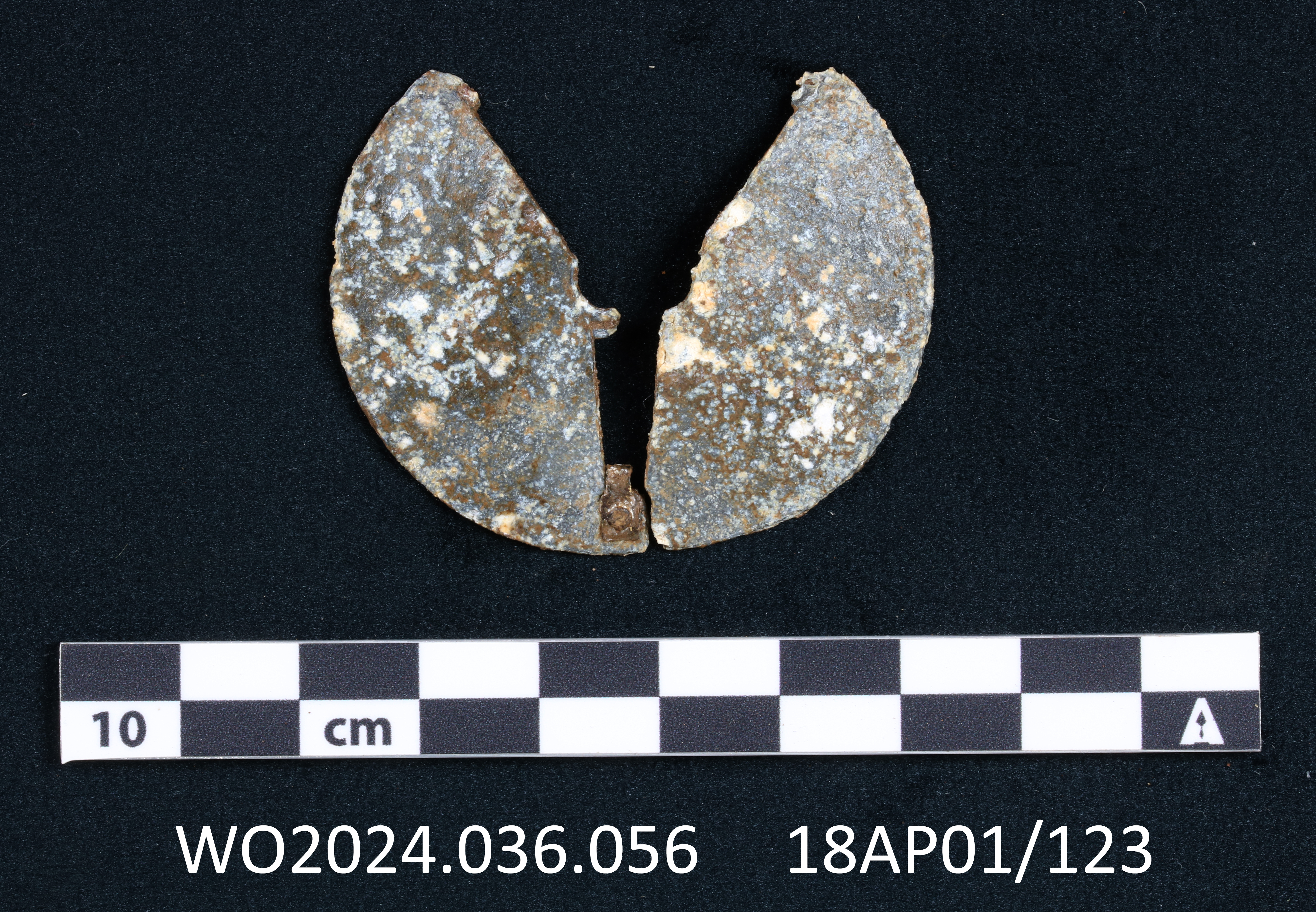 Figure 1 Incomplete aluminum disc with rough surface broken into two. A Y-shaped gap tapers to
the center of the disc and extends in a slot next to a small copper alloy stub adjacent to
the slot. The sides of the Y-shaped gap shows cut indentations that might indicate
similar slots on the edge of the Y. A photo scale shows the artifact is less than 5
centimeters in diameter.