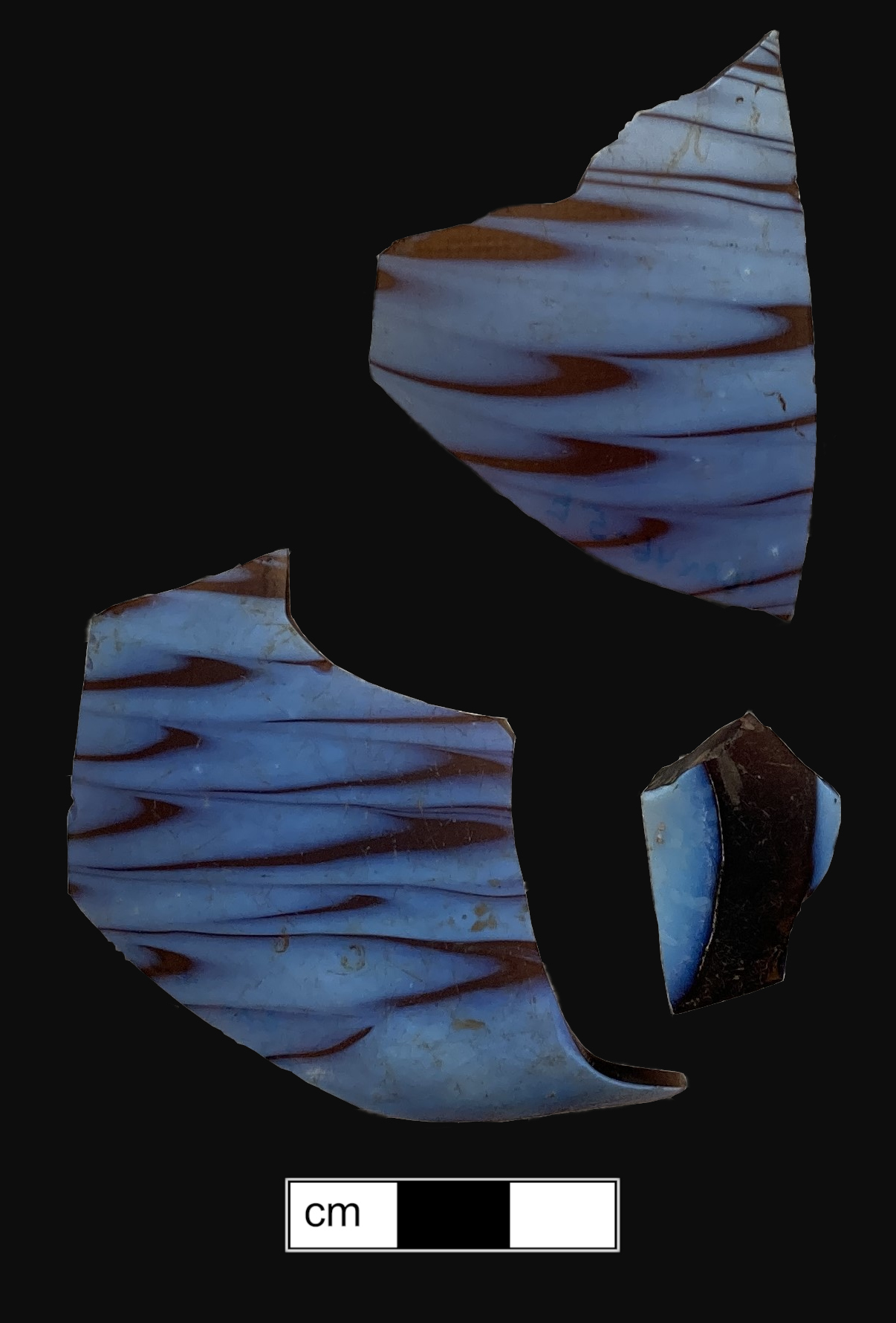 Three fragments of amber glass with blue combed decoration on a black background with scale bar in centimeters.