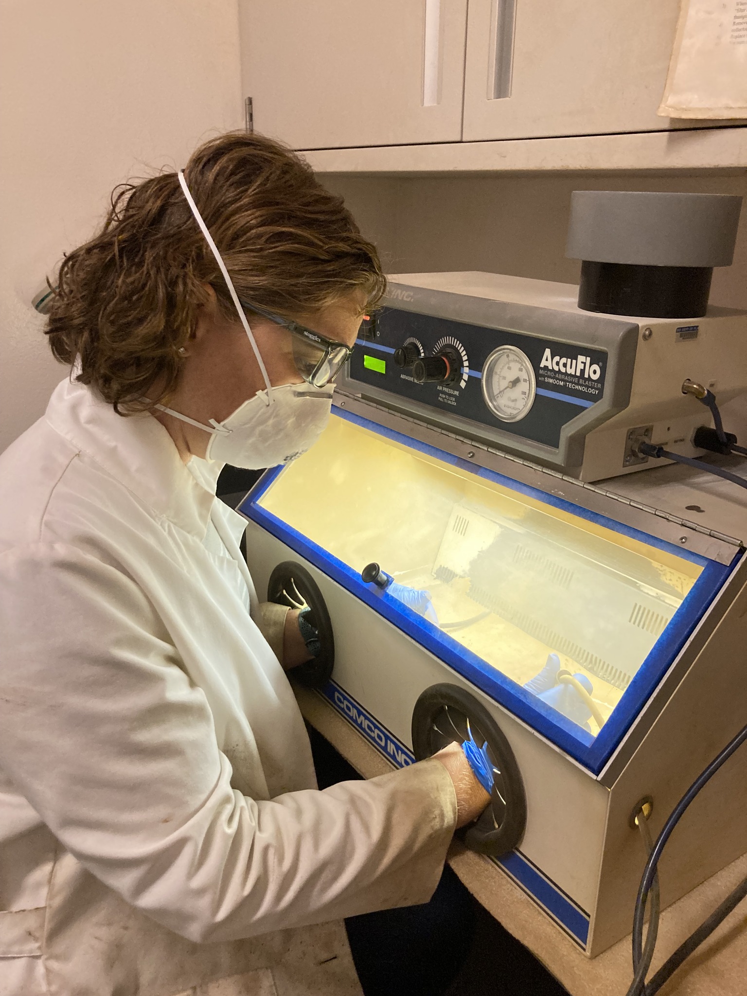 A conservator in a white lab coat, safety glasses, and dust mask operates a micro-abrasive blaster, working through the cabinet's hand ports to clean or treat an artifact inside the illuminated enclosure.