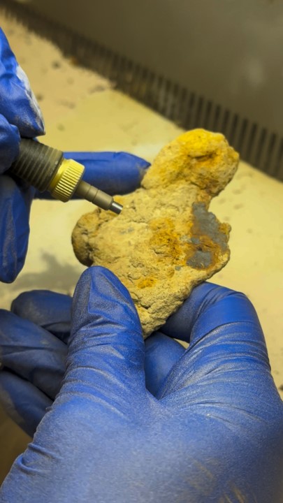 Gloved hands hold a corroded metal artifact while a micro-abrasive blaster nozzle is directed at its surface, removing orange and yellow corrosion products to reveal the underlying material.