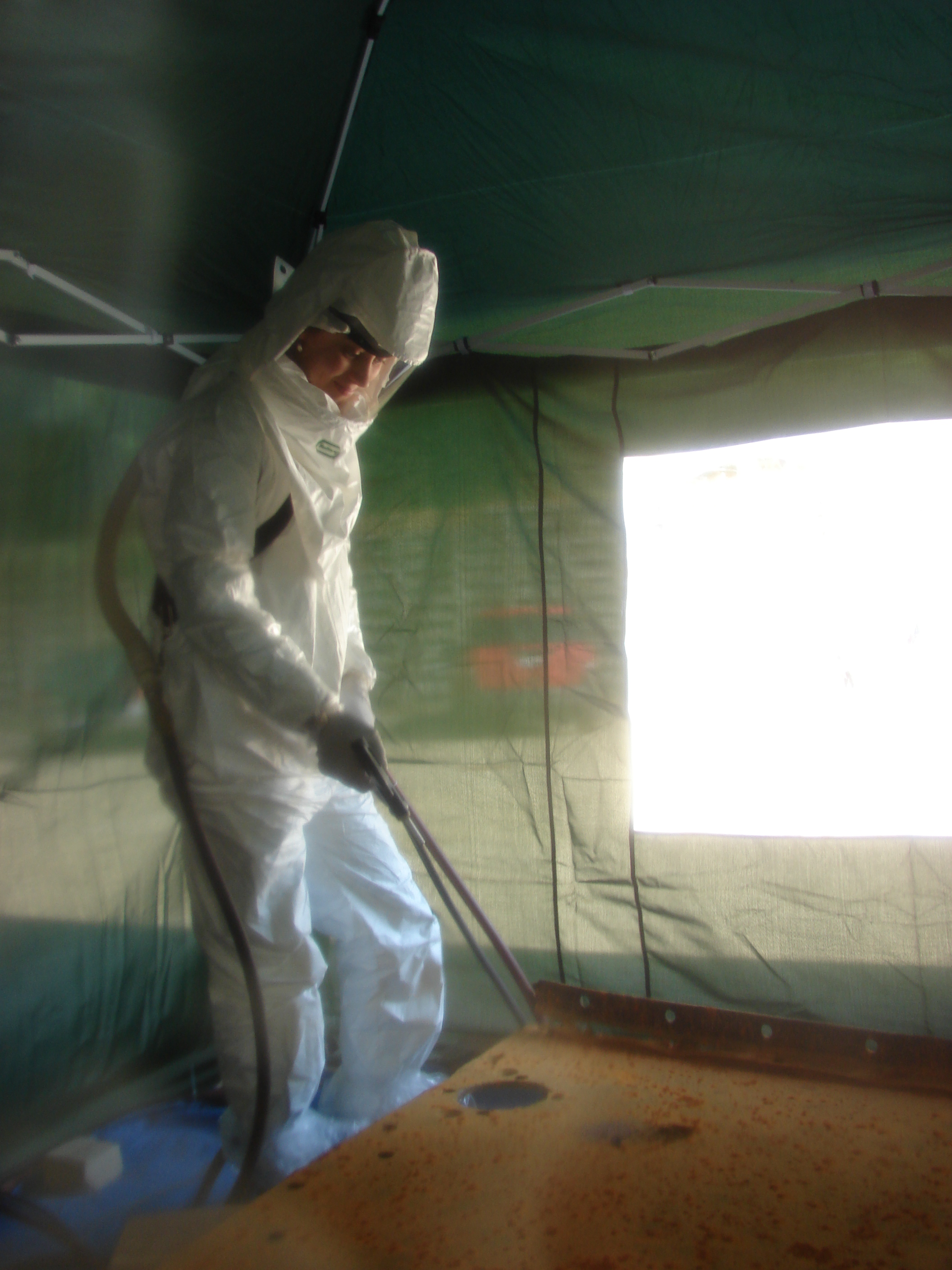 A conservator in a full Tyvek suit and hood directs an air abrasive lance at a large corroded metal object inside a containment tent during large-scale conservation treatment.