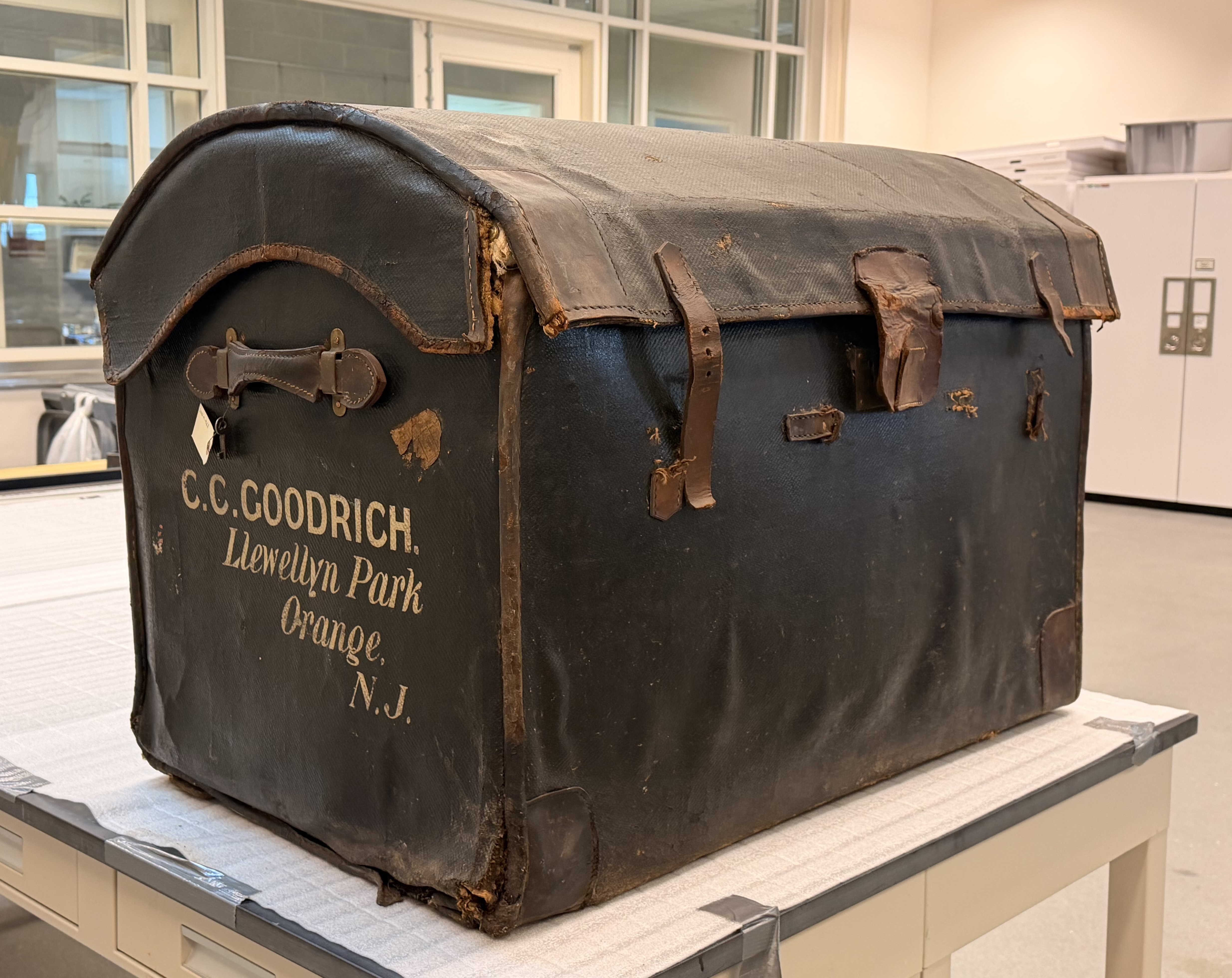 Black travel trunk belonging to C.C. Goodrich, featuring exterior straps and the inscription 