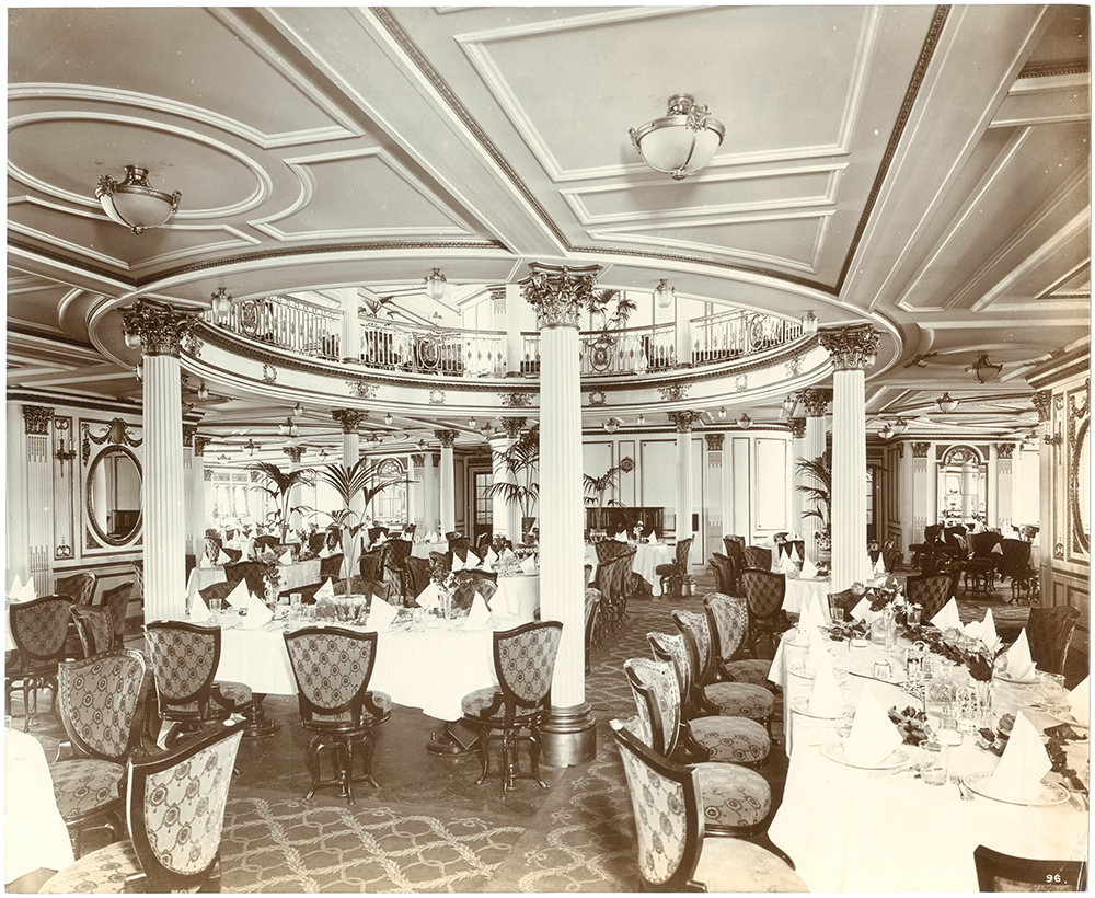 The interior of the first-class dining saloon on the RMS Lusitania, showcasing an opulent and elegantly designed space. The room features a high, intricately designed ceiling with decorative moldings and several ornate light fixtures. White tables are set with fine linens, glassware, and floral arrangements, surrounded by plush, patterned chairs with armrests.