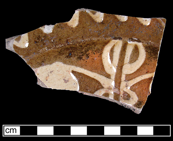 North Midlands reverse slip decorated dish.  Decorated in white slip in possible floral pattern. Interior of vessel covered with red slip and white slip trailed decoration applied over red slip. Glazed interior and unglazed brown slip dipped exterior - Collected by George L. Miller in 1986 in Staffordshire, England.
